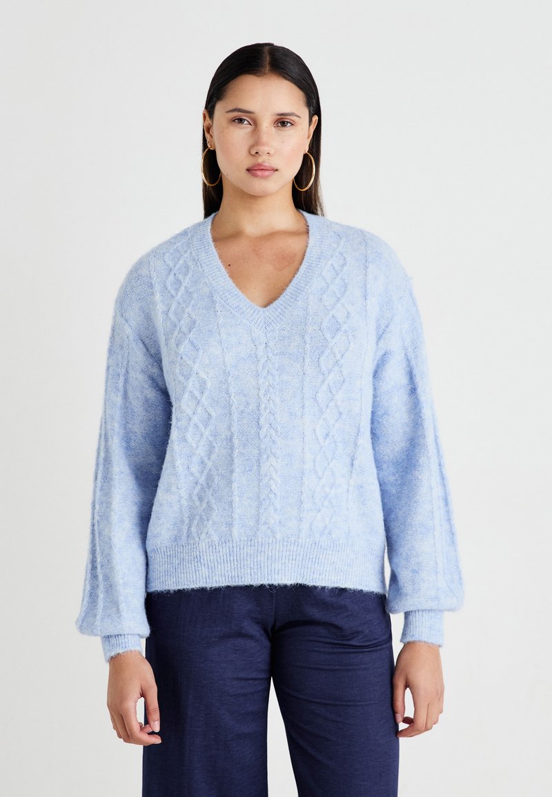 Hellblau gestrickter Pullover mit V-Ausschnitt, Zopfmuster, langen Puffärmeln und geripptem Saum. Weiche Textur. Kombiniert mit dunklen Hosen.