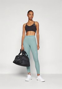 Fekete sportmelltartót, tengerkék Puma leggingset és fehér tornacipőt viselő nő, aki egy fekete sporttáskával áll egy világos, egyszínű háttér előtt.