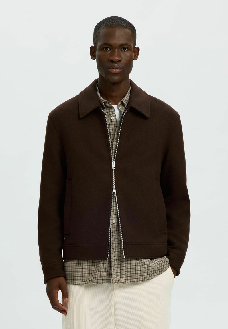Jeune homme portant une veste zippée marron foncé par-dessus une chemise à carreaux et un pantalon clair, debout devant un fond uni.
