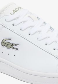 Sneaker bianca in pelle con finitura testurizzata, caratterizzata da un logo di coccodrillo ricamato verde e lacci bianchi. Design minimalista, perforazioni sobrie.
