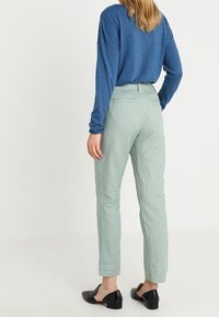 Pantalons vert clair effilés avec une finition texturée et des poches latérales, assortis à un haut bleu à manches longues et des chaussures plates noires.