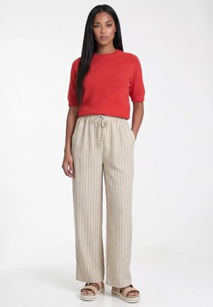 Kobieta stojąca, ubrana w czerwoną krótką bluzkę, beżowe szerokie spodnie w cienkie paski ze sznurkiem oraz beżowe sandały na platformie, na białym tle.