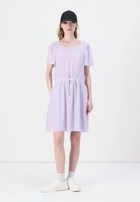 Lilac pullover-mekko, jossa on lyhyet hihat, joustava vyötärö, nyöri, sivutaskut ja sileä rakenne, yhdistettynä valkoisiin lenkkareihin ja mustaan lippalakkiin.