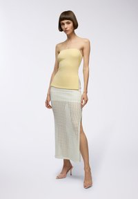 Top giallo pallido senza spalline abbinato a una lunga gonna bianca in crochet con uno spacco laterale. La modella indossa tacchi trasparenti, che evidenziano un design minimalista.