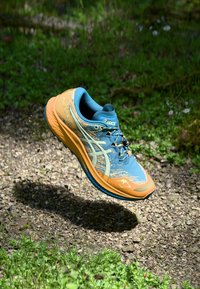 Blauwe en oranje ASICS hardloopschoen met een gestructureerde bovenkant, gebogen silhouette en zichtbaar logo, zwevend boven een grindoppervlak met groen.