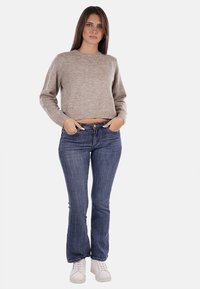 Maglione corto marrone chiaro con decorazioni, abbinato a jeans a zampa d'elefante blu e sneakers bianche. La modella posa con le mani sui fianchi.