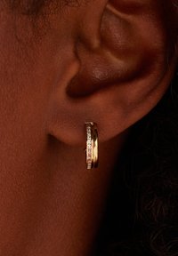 Gouden hoopoorbel met een gladde oppervlakte en een glinsterende rhinestone-accent aan de ene kant, bevestigd aan een close-up van een oor.