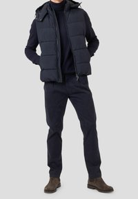 Chaleco acolchado azul marino con cuello alto y capucha, llevado sobre una camisa de manga larga oscura, combinado con pantalones oscuros a juego y botines marrones.