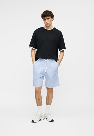 Νέος άνδρας που στέκεται με τα χέρια στις τσέπες, φοράει μαύρο T-shirt, ανοιχτόγαλανες σόρτς με κορδόνι, λευκές κάλτσες και λευκά αθλητικά παπούτσια μπροστά σε λευκό φόντο.