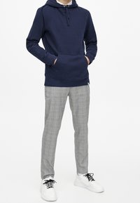 Mörkblå hoodie med framficka och dragsko, kombinerad med grå rutiga byxor och vita sneakers. Slätt tyg och avslappnad passform.