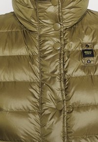 Chaleco acolchado de color verde oliva con un cuello alto, que presenta grandes botones redondos y un diseño acolchado. Incluye una etiqueta de la marca en el lateral.