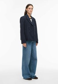 Femme portant un blazer croisé bleu marine, un jean bleu à jambes larges et des chaussures noires, debout devant un fond blanc.