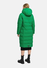 Abrigo largo tipo plumas en verde brillante. Cuenta con capucha, forma cuadrada, costuras horizontales y un parche con el logo en la parte inferior. Combinado con botas negras.