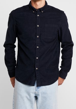 Homme portant une chemise en velours côtelé bleu marine foncé avec une poche poitrine et un jean bleu clair, debout les mains dans les poches.