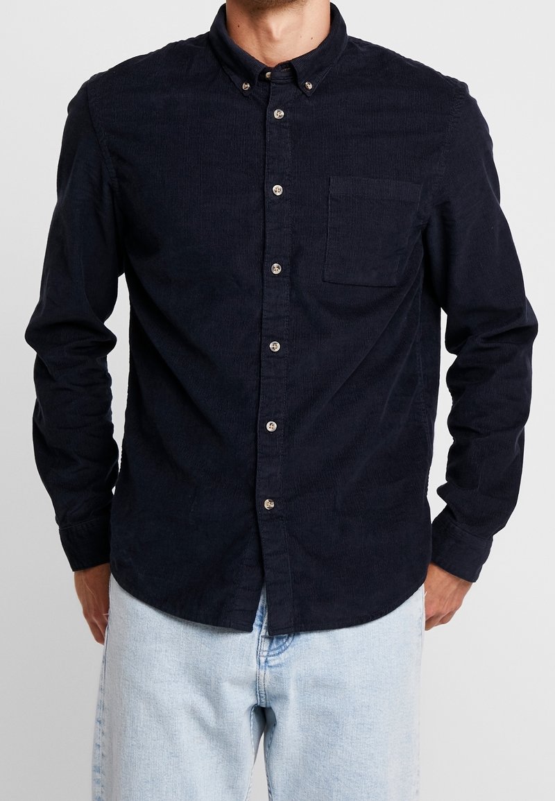 Homme portant une chemise en velours côtelé bleu marine foncé avec une poche poitrine et un jean bleu clair, debout les mains dans les poches.