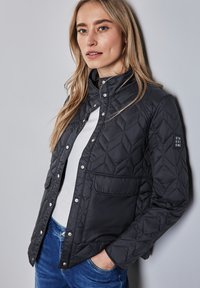 Schwarze gesteppten Jacke mit Stehkragen, Druckknöpfen und einer Pattentasche. Mit Rautenmuster-Stitching und Logo auf dem Ärmel.
