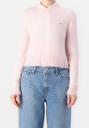 Person trägt einen hellrosa, flauschigen Strickpullover mit geripptem Saum und Bündchen, kombiniert mit hoch taillierten blauen Jeans.