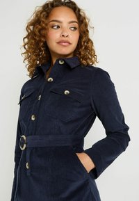 Femme aux cheveux bouclés portant une veste en velours côtelé bleu marine ceinturée avec des poches poitrine boutonnées, la main dans la poche latérale, regardant sur le côté.