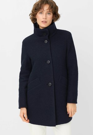 Woman wearing a dark navy high-collar coat with four front buttons and two slanted pockets, paired with white pants.