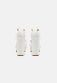 Zapatos de tacón alto blancos con punta redonda y correa en el tobillo. Fabricados con un tejido suave, cuentan con un tacón grueso y una suela marrón sutil.