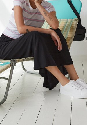 Personne tatouée portant un chemisier rayé et une jupe noire, assise sur une chaise longue tressée, avec des baskets blanches et un sac noir.