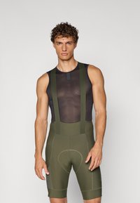 Bibshorts i oliven grønn med svart mesh-top. Har brede stropper, polstret sete og en tettsittende passform. Materialet ser glatt og elastisk ut.