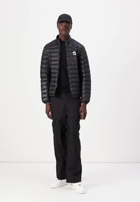 Schwarze Pufferjacke mit horizontalen Nähten und kleinem Logo, kombiniert mit schwarzen Cargo-Hosen und weißen Turnschuhen sowie einer passenden Mütze.