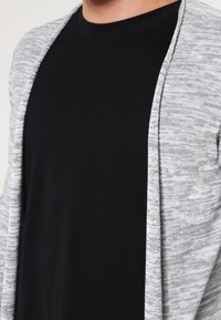 Lång grå cardigan med melerad textur, öppen front och långa ärmar, lagd över en enfärgad svart t-shirt.