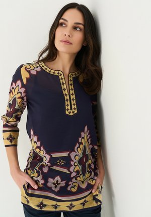 Femme aux longs cheveux foncés portant une tunique bleu marine avec des motifs ornés jaunes et bordeaux, debout contre un mur blanc, les mains dans les poches.