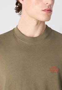 Olivově zelené bavlněné tričko s žebrovaným výstřihem a červeným vyšitým logem na hrudi. Hladká textura s přiléhavým střihem.