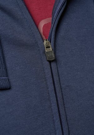 Jūras spēku zipveida džemperis ar teksturētu audumu un gludu rāvējslēdzēju, kārtots virs bordo krāsas krekla ar redzamu grafisku rakstu.