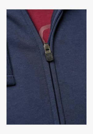 Navy zip hoodie met een textielstructuur en een gladde rits, laag over een bordeaux shirt met een zichtbaar grafisch patroon.