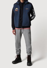 Gillet in pile blu con dettagli neri, zip frontale e toppe; abbinato a pantaloni sportivi grigi e stivali neri con lacci. Design semplice, vestibilità rilassata.