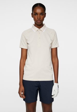 Polo beige con colletto bianco, maniche corte e chiusura a bottoni; abbinato a pantaloni corti blu e un guanto da golf bianco su una mano.