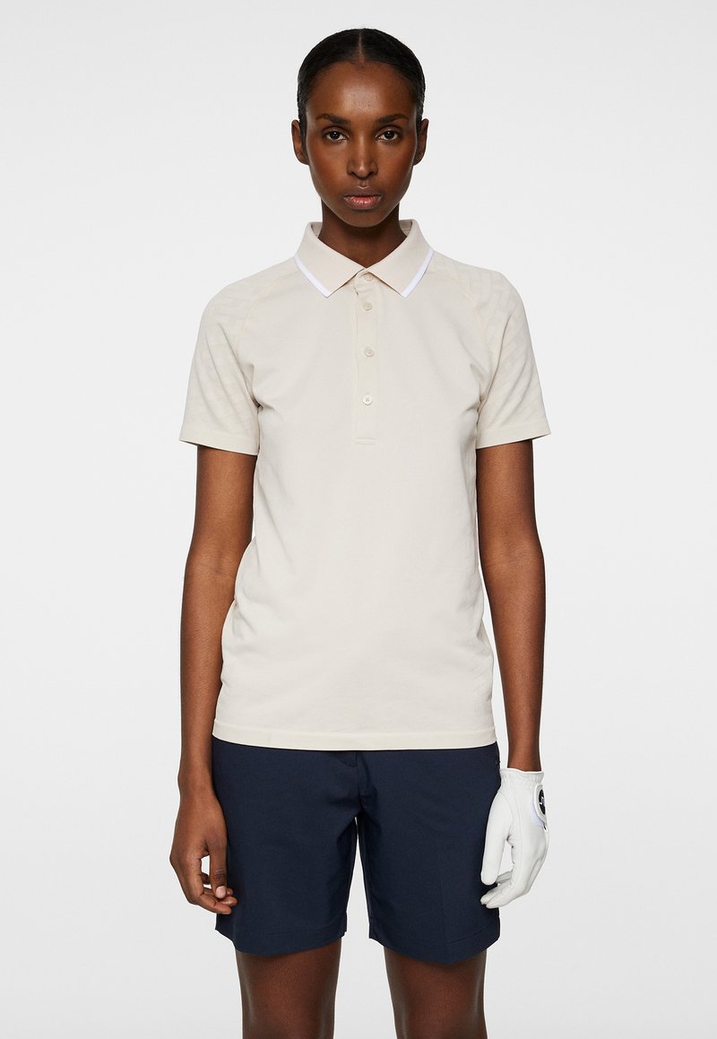 Beige pikétröja med vit krage, korta ärmar och knäppning fram; kombinerad med marinblå shorts och en vit golfhandske på ena handen.