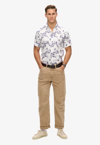 Camisa blanca de manga corta con un patrón floral azul, combinada con chinos beige y zapatillas color crema. Se puede ver un cinturón y un reloj marrones.