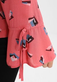 Röd blus med utsvängda ärmar som har geometriska mönster i blått, svart och vitt. Inkluderar en knytdetalj vid ärmarnas fåll.