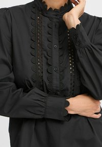 Black blouse with high collar, scalloped edges, and decorative stitching. Features button closure and gathered sleeves for a textured look.