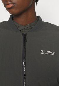 Giacca leggera grigia con colletto a coste, zip frontale e logo "new balance" bianco. Presenta una texture liscia e affusolata.