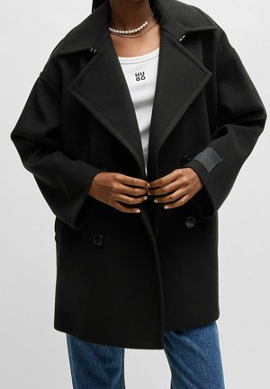 Osoba ubrana w oversize'owy czarny dwurzędowy płaszcz, biały top z napisem „HUGO”, niebieskie dżinsy i perłowy naszyjnik, zapinająca guziki płaszcza.