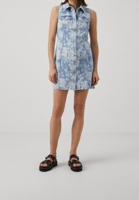 Denim dress in light blue with white floral pattern, sleeveless design, button-down front, and two chest pockets; paired with black sandals.