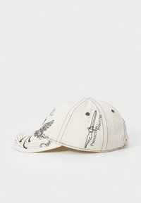 Cap in light beige fabric featuring black graphic prints, including an eagle and stylized text. Curved visor with five panels and stitching details.