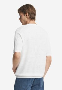 Kortärmad, räfflad vit tröja med rund halsringning och mjuk textur, kombinerad med blå denimjeans. Designen är enkel och ren.