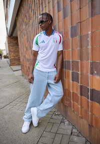 Biele talianske futbalové tričko so zelenými, červenými a modrými akcentmi; svetlomodré voľné džínsy; biele tenisky; čierne slnečné okuliare; stojí opretý o tehlovú stenu.
