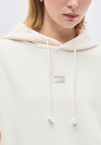 Γυναίκα που φοράει ένα εκρού φούτερ με κορδόνια και ένα μικρό ορθογώνιο λογότυπο "Tommy Jeans" στο κέντρο του στήθους, ορατή από τους ώμους μέχρι τα χείλη.