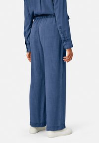 Une personne portant un pantalon bleu ample et un haut à manches longues assorti, se tenant debout avec les mains détendues sur les côtés, des baskets blanches visibles.