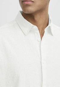 Camisa de lino blanca con cuello abotonado y tapeta frontal, que muestra una tela suave y texturizada. No se observan patrones ni acentos.