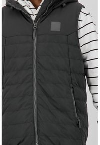 Zwarte puffer bodywarmer met horizontaal geborduurd patroon, voorrits en zijzakken, gedragen over een zwart-wit gestreept langshirt.