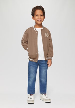 Braune Bomberjacke mit Reißverschluss und weißen Streifen am Kragen und an den Bündchen, getragen über einem weißen T-Shirt, kombiniert mit blauen Jeans und weißen Sneakers.