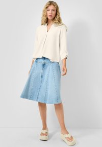 Mujer rubia con una blusa blanca suelta, falda de mezclilla azul claro en línea A y sandalias beige con plataforma, sobre un fondo claro y simple.